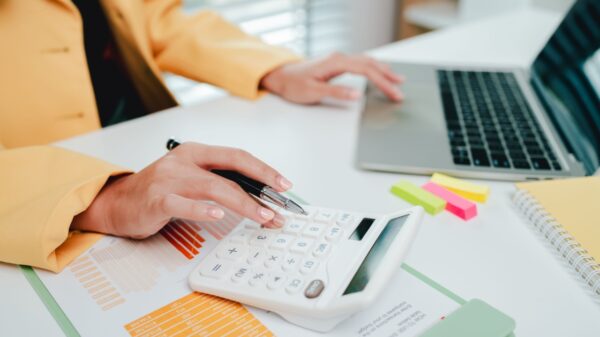 par de mãos feminina onde a mão esquerda está em um notebook e a mão direita está em uma calculadora e o ambiente é um escritório