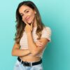 mulher no centro da imagem sorrindo e com a mão na altura do queixo em fundo azul