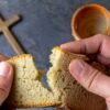 imagem de um copo e uma cruz em cima de uma mesa e em primeiro plano uma mão masculino dividindo um pão