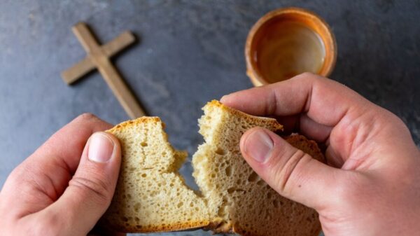 imagem de um copo e uma cruz em cima de uma mesa e em primeiro plano uma mão masculino dividindo um pão