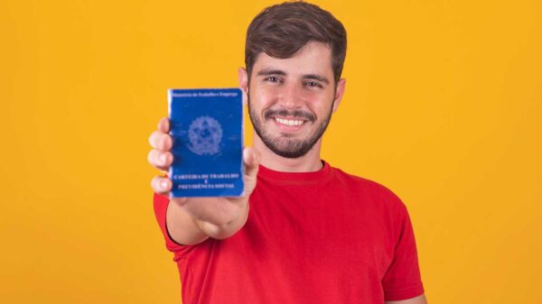 homem com camisa vermelha e segurando uma carteira de trabalho na mão direita em fundo amarelo