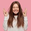 mulher jovem sorrindo com os dedos cruzados em fundo rosa