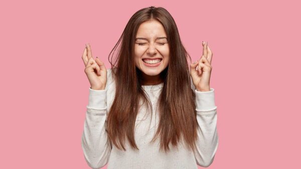 mulher jovem sorrindo com os dedos cruzados em fundo rosa