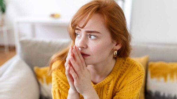 mulher ruiva sentada em um sofá com as mãos juntas e olhando para o lado com expressão de preocupada