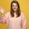 mulher com um sorriso no rosto e fazendo sinal de tchau com a mão direita em fundo amarelo