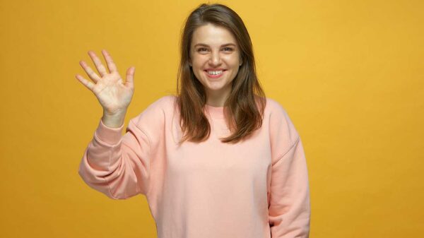 mulher com um sorriso no rosto e fazendo sinal de tchau com a mão direita em fundo amarelo