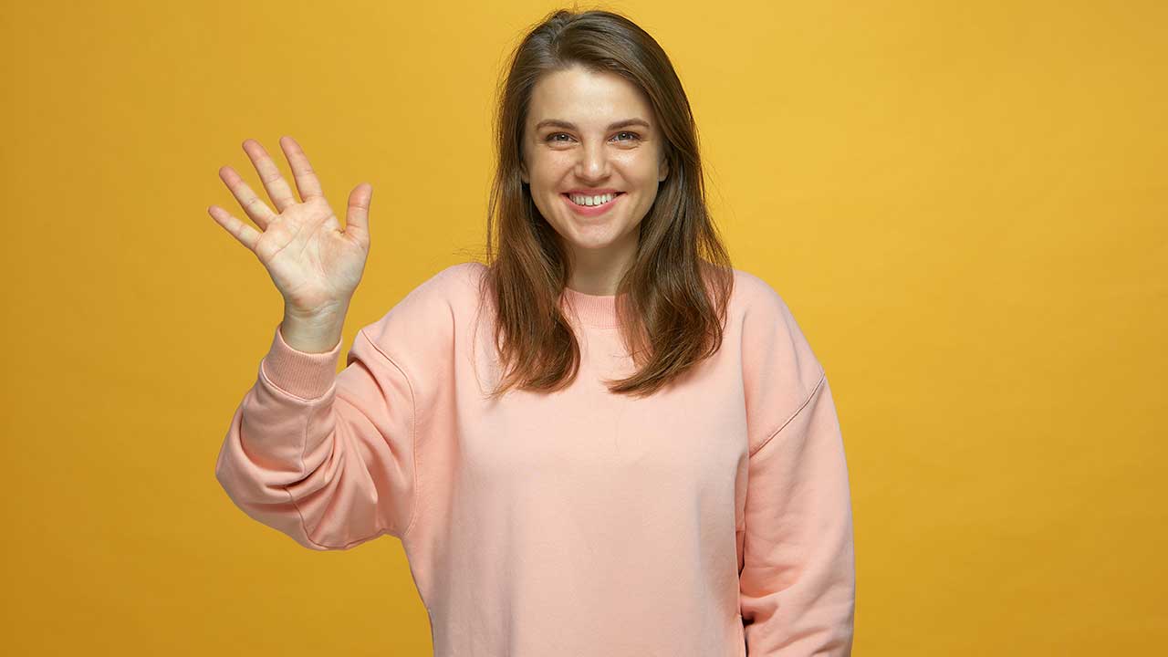 mulher com um sorriso no rosto e fazendo sinal de tchau com a mão direita em fundo amarelo