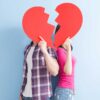casal heterossexual de costas uma para o outro e segurando um coração vermelho grande e partido tampando os rostos em fundo azul
