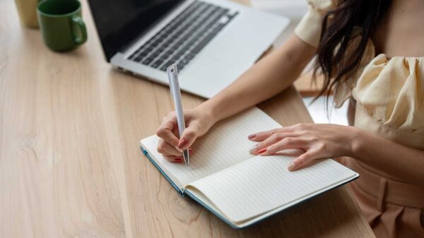 menina escrevendo em um caderno com linhas em cima de uma mesa de madeira com um notebook do lado