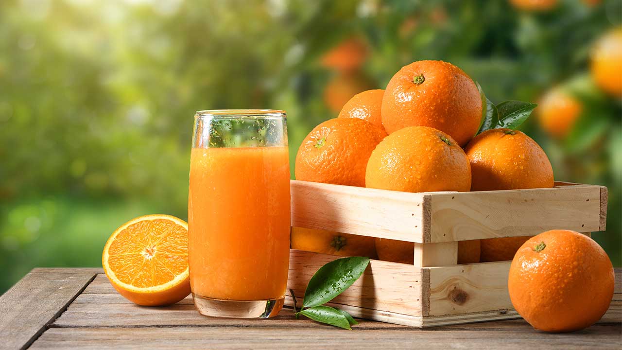 mesa de madeira com uma caixa de madeira cheia de laranjas e um copo de vidro com suco de laranja e meia laranja do lado em fundo desfocado com pés de laranja