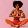 mulher sentada com roupas de ginástica sentada em posição de yoga em fundo rosa
