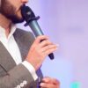 homem com roupa social e um microfone na mão aparentemente falando em um evento