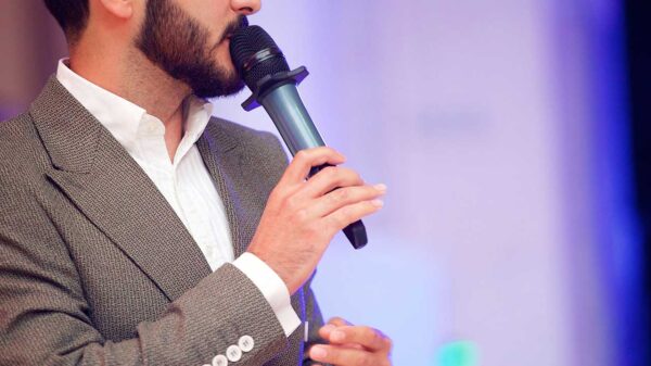 homem com roupa social e um microfone na mão aparentemente falando em um evento