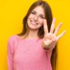mulher jovem sorrindo fazendo o número quatro com os dedos em fundo amarelo