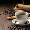 Café quente em uma xícara de café branca e muitos grãos de café colocados ao redor e açúcar em uma mesa de madeira em um ambiente quente e leve, em fundo de madeira escuro