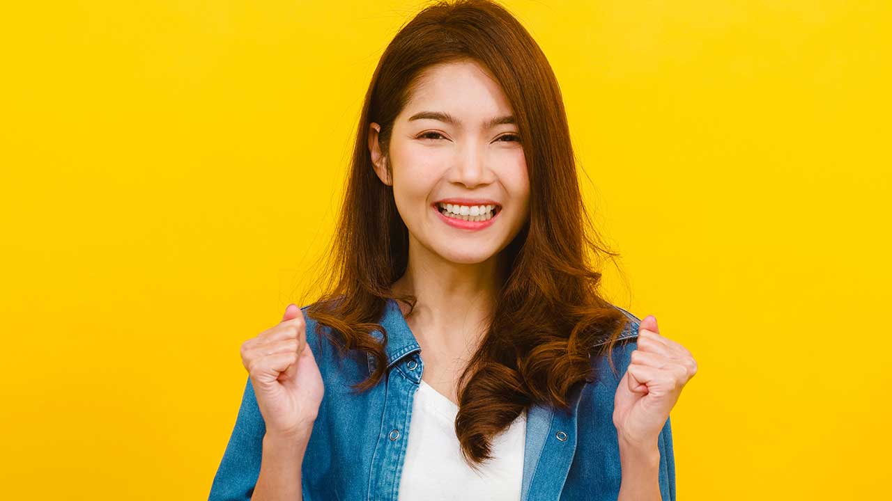 mulher jovem com traços asiáticos em expressão corporal de comemoração em fundo amarelo