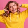 mulher sorrindo com as mãos na altura de cabeça e moletom amarelo em fundo rosa