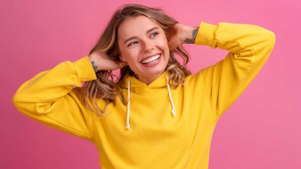 mulher sorrindo com as mãos na altura de cabeça e moletom amarelo em fundo rosa