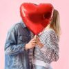 casal heterossexual se beijando com um balão em formato de coração vermelho tampando os rostos em fundo rosa