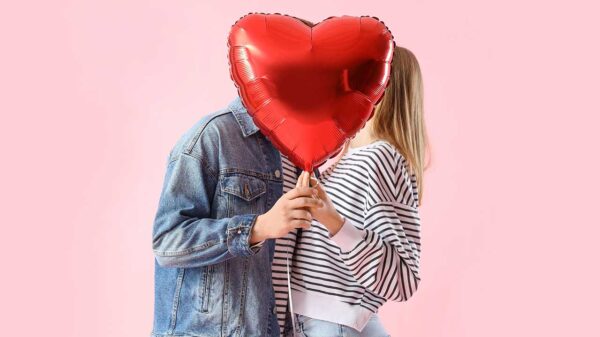 casal heterossexual se beijando com um balão em formato de coração vermelho tampando os rostos em fundo rosa