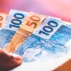 fotografia de uma mão segurando duas notas de 50 reais e duas notas de 100 reais em um fundo desfocado