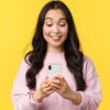 mulher olhando sorrindo para um celular que está em suas mãos em fundo amarelo