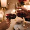 mãos de um casal brindando uma taça de vinho cada um em um fundo romântico a luz de velas