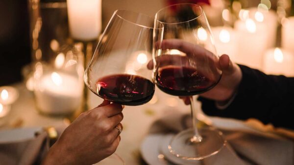 mãos de um casal brindando uma taça de vinho cada um em um fundo romântico a luz de velas