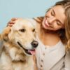 mulher jovem sorrindo e fazendo carinho em um cachorro grande de pelos loiros em fundo azul