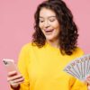 mulher segurando um celular na mão direita e um leque de euros na mão esquerda em fundo rosa