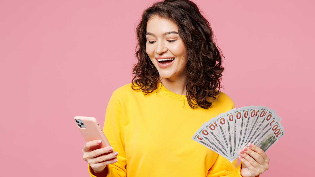 mulher segurando um celular na mão direita e um leque de euros na mão esquerda em fundo rosa
