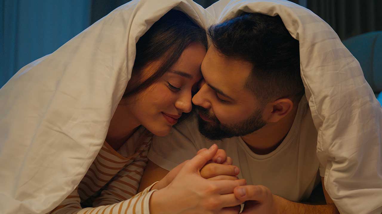 casal formado por um homem e uma mulher deitado embaixo de cobertores vestidos e de mãos dadas