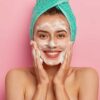 mulher jovem sorrindo com uma toalha verde enrolada na cabeça e espuma no rosto em fundo rosa
