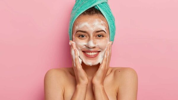 mulher jovem sorrindo com uma toalha verde enrolada na cabeça e espuma no rosto em fundo rosa