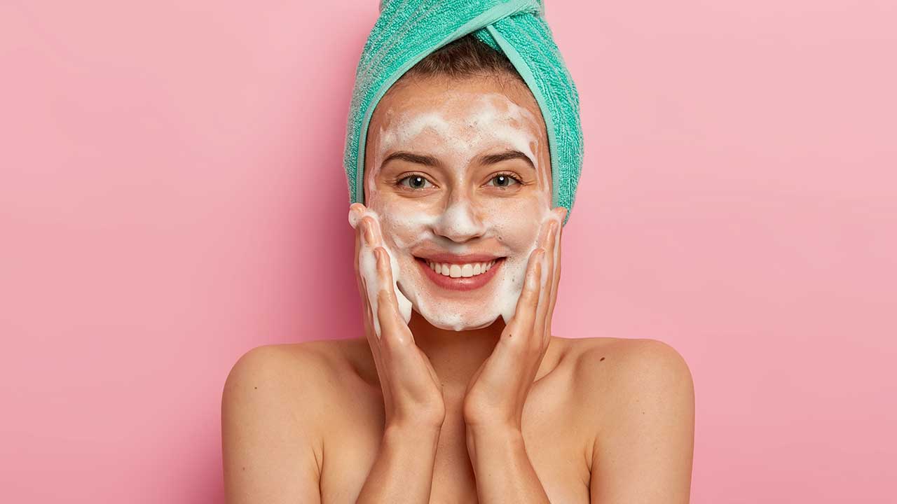 mulher jovem sorrindo com uma toalha verde enrolada na cabeça e espuma no rosto em fundo rosa