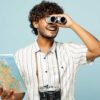 homem no centro da imagem segurando um mapa na mão direita e um binóculos na esquerda com uma câmera fotográfica no pescoço em fundo azul