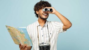 homem no centro da imagem segurando um mapa na mão direita e um binóculos na esquerda com uma câmera fotográfica no pescoço em fundo azul