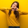 mulher jovem com moletom amarelo fazendo um coração com a mão em fundo amarelo