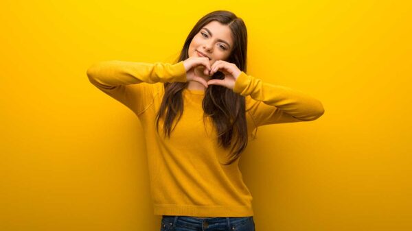mulher jovem com moletom amarelo fazendo um coração com a mão em fundo amarelo