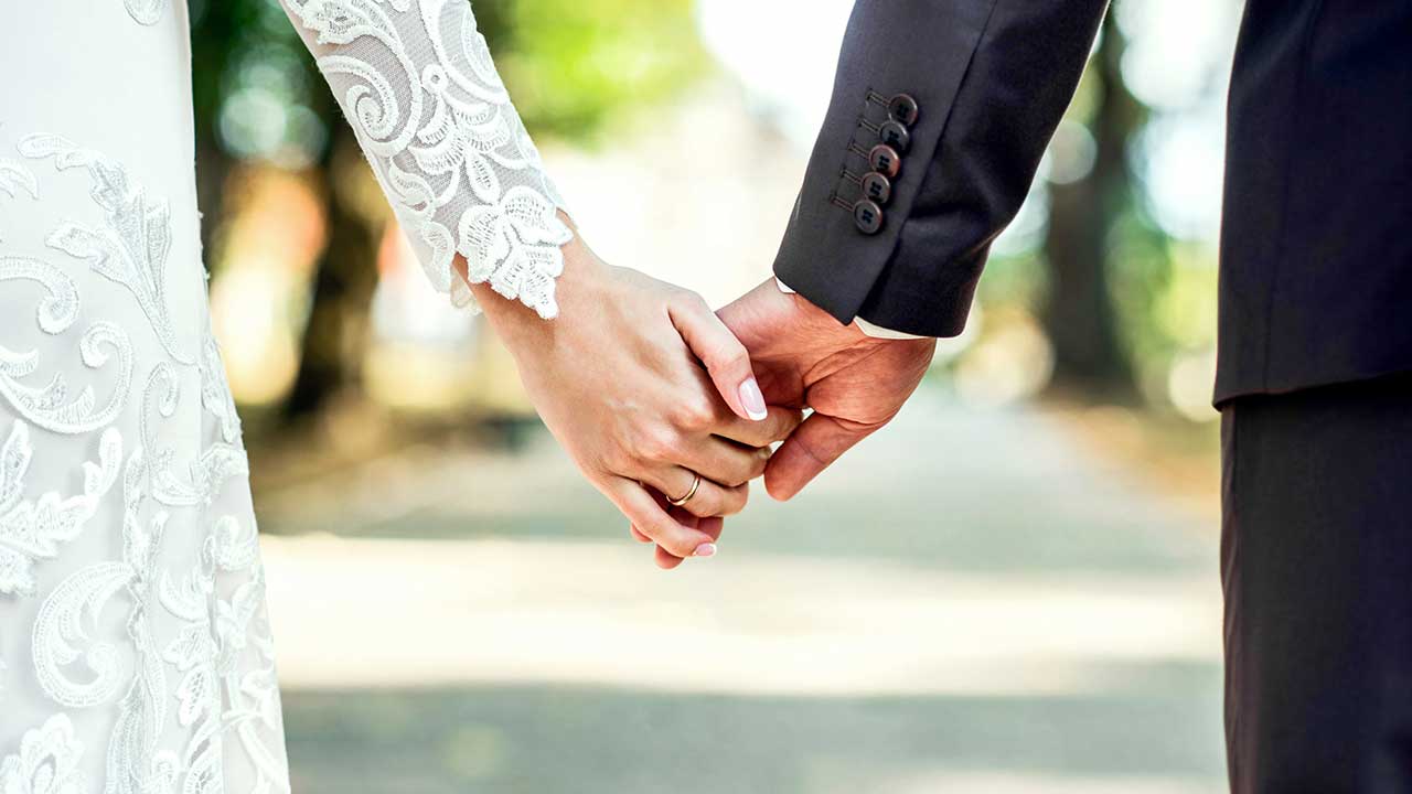 imagem de mãos dadas em um fundo aberto e desfocado onde ela usava vestido de noiva e ele terno