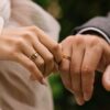 fotografia de mãos de um casal se tocando e com alianças douradas no dedo anelar e eles estão com roupa de noiva e noivo