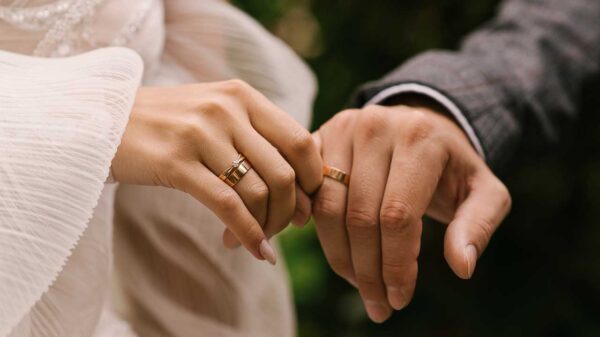 fotografia de mãos de um casal se tocando e com alianças douradas no dedo anelar e eles estão com roupa de noiva e noivo