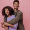 casal formato por um homem e uma mulher abraçados e sorrindo em fundo rosa