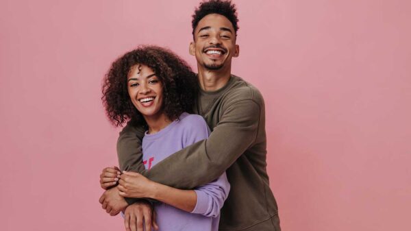 casal formato por um homem e uma mulher abraçados e sorrindo em fundo rosa
