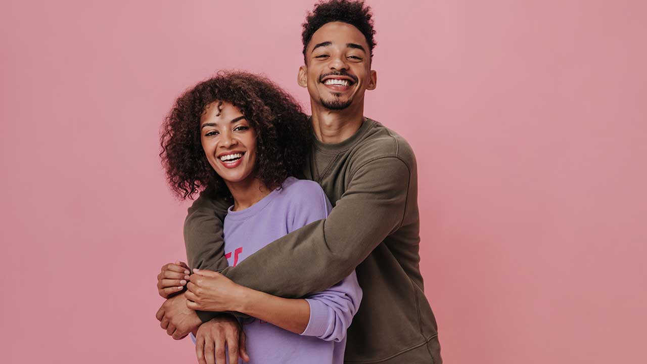 casal formato por um homem e uma mulher abraçados e sorrindo em fundo rosa