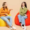 duas amigas sentadas cada uma em um puff vermelho conversando em fundo salmão