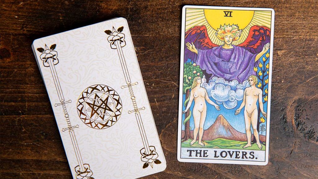 carta do tarot os enamorados ao lado de um deck branco de tarot em cima de uma mesa de madeira