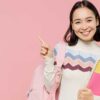 mulher segurando caderno e uma mochila nas costas em fundo rosa